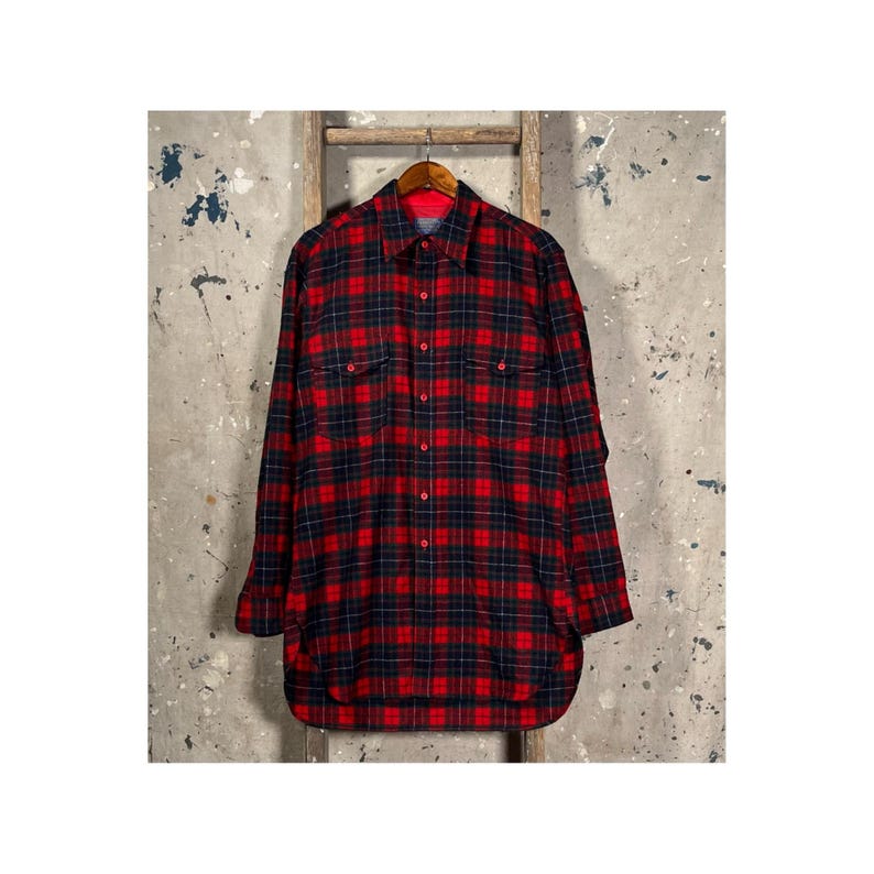 Puede incluir: Una camisa a cuadros rojos y negros con botones, mangas largas y dos bolsillos en el pecho. La camisa est&aacute; colgada en una percha de madera sobre un fondo neutro.