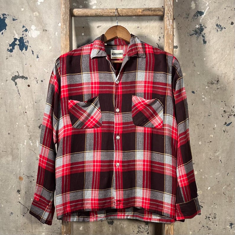 May include: A long-sleeved, button-down plaid shirt in red, black, and gray. The shirt has two chest pockets and is hanging on a wooden hanger. The shirt's collar is open, and the fabric has a soft, slightly worn appearance.