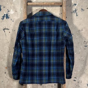 May include: A long-sleeved plaid shirt in shades of blue, green, and navy hangs on a wooden hanger. The shirt features a classic button-down collar and a traditional plaid pattern. The fabric appears to be a medium-weight material, suitable for cooler weather.