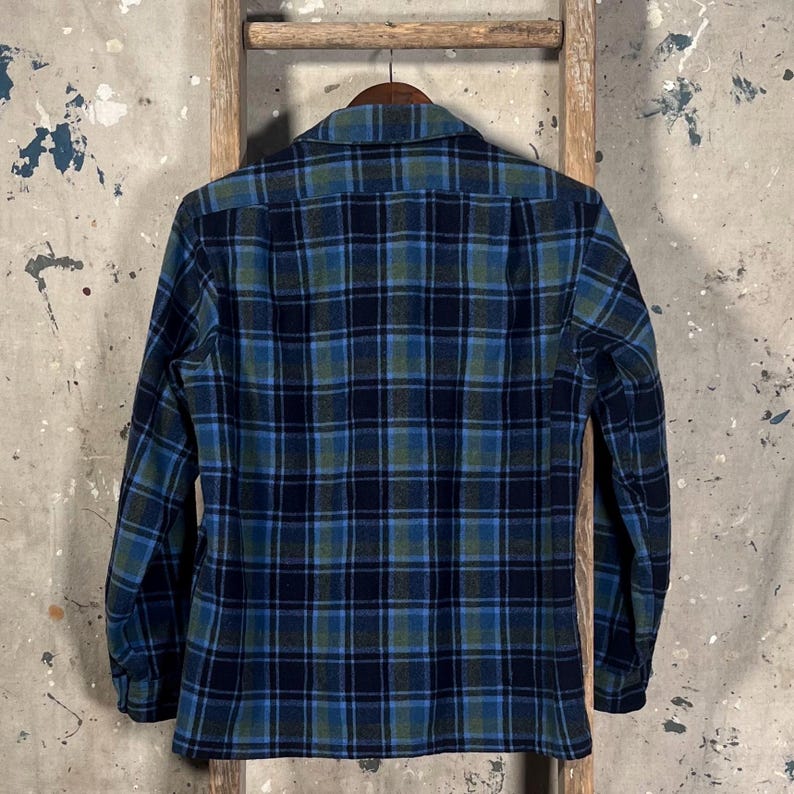 May include: A long-sleeved plaid shirt in shades of blue, green, and navy hangs on a wooden hanger. The shirt features a classic button-down collar and a traditional plaid pattern. The fabric appears to be a medium-weight material, suitable for cooler weather.