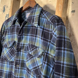 May include: A blue, black, and green plaid flannel shirt with two chest pockets and black buttons. The shirt is hanging on a wooden hanger against a rustic background. The shirt has a classic, button-down design.