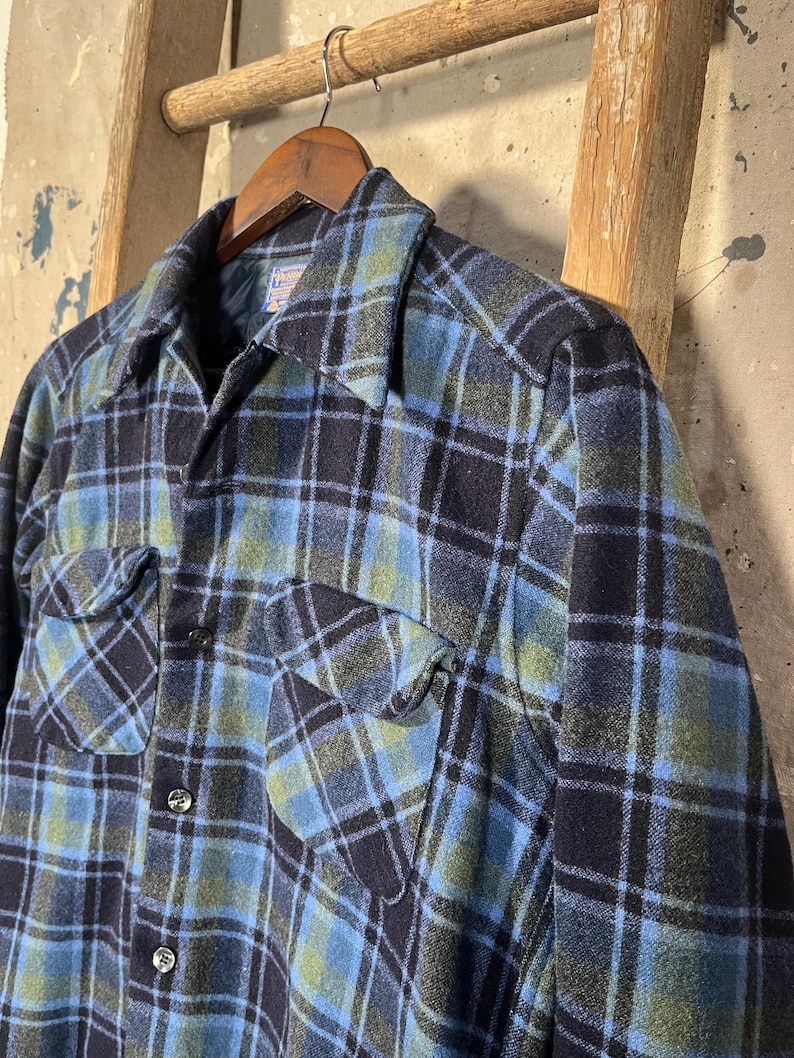 May include: A blue, black, and green plaid flannel shirt with two chest pockets and black buttons. The shirt is hanging on a wooden hanger against a rustic background. The shirt has a classic, button-down design.