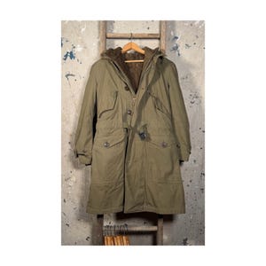May include: An olive green winter coat with a hood and a brown fur lining. The coat has a belt, pockets, and buttons. It is hanging on a wooden ladder against a distressed concrete wall.