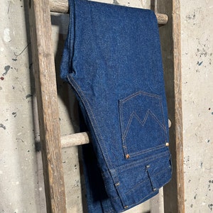 Puede incluir: Un par de pantalones vaqueros de mezclilla azul oscuro colgados de una escalera de madera. Los vaqueros tienen un estilo cl&aacute;sico de pierna recta y un dise&ntilde;o tradicional de cinco bolsillos.