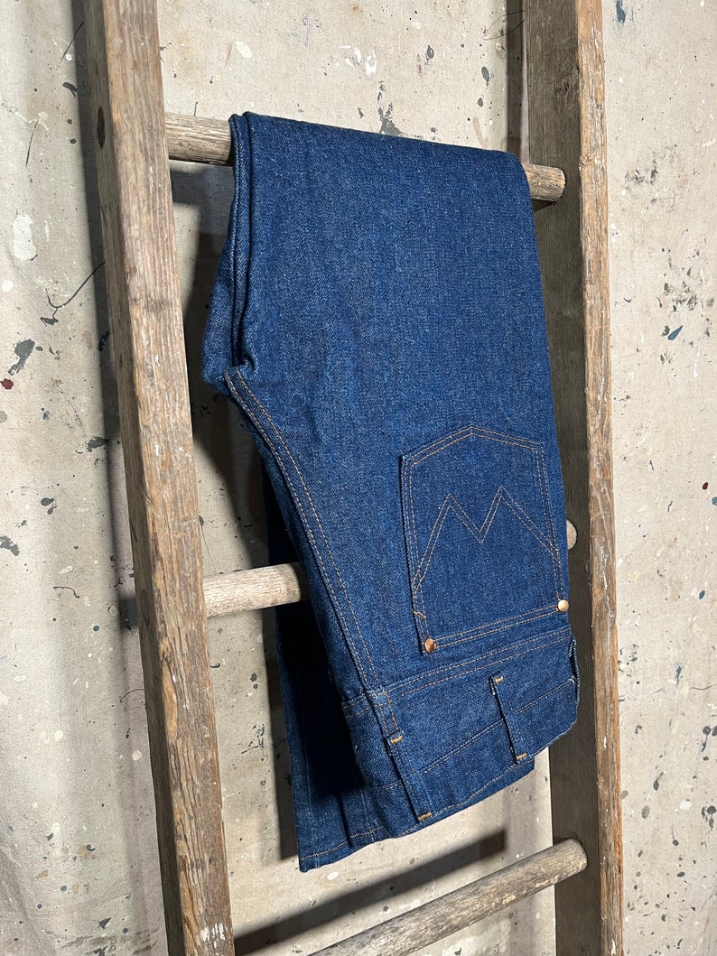 Puede incluir: Un par de pantalones vaqueros de mezclilla azul oscuro colgados de una escalera de madera. Los vaqueros tienen un estilo cl&aacute;sico de pierna recta y un dise&ntilde;o tradicional de cinco bolsillos.