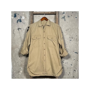 Puede incluir: Una camisa beige de manga larga con botones y dos bolsillos de solapa en la parte delantera. La camisa tiene un corte holgado y está hecha de una tela ligera.