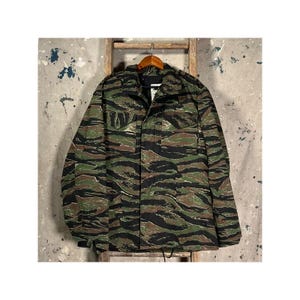 May include: A long-sleeve jacket featuring a tiger stripe camouflage pattern in shades of black, olive green, and brown. The jacket includes a fold-down collar, a full-length front zipper closure, and two chest pockets with buttoned flaps.