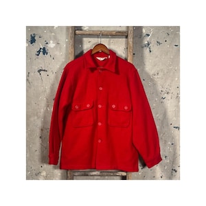 May include: A bright red button-down shirt with two chest pockets and a collar. The shirt is made of a thick, warm material, perfect for cooler weather. The shirt is hanging on a wooden hanger.