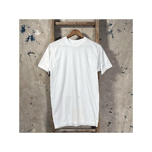 Puede incluir: Una camiseta blanca lisa con cuello redondo cuelga de una percha de madera. La camisa está hecha de un material suave y opaco. El fondo es una escalera de madera desgastada contra una pared moteada.