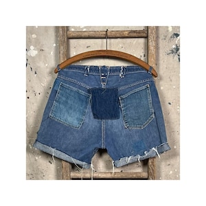 Puede incluir: Pantalones cortos vaqueros azules con bolsillos parcheados y dobladillo vuelto. Los pantalones cortos cuelgan de una percha de madera sobre un fondo rústico. Los pantalones cortos tienen un aspecto único y desgastado.