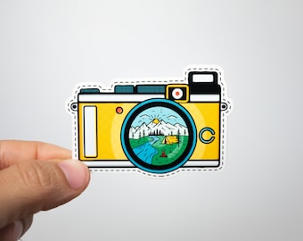 May include: A yellow and white sticker in the shape of a vintage camera. The camera lens displays a scenic landscape with mountains, a river, and a tent. The sticker is held by a hand.