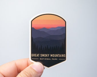 May include: A sticker featuring a landscape of the Great Smoky Mountains National Park. The design includes a sunset with orange and red hues over layered blue mountains and a forest silhouette. The text "GREAT SMOKY MOUNTAINS NATIONAL PARK" is displayed.
