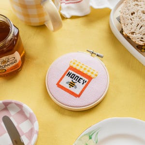 May include: A pink and white cross-stitch embroidery hoop with a design of a honey jar with a bee and the word "honey" on it.