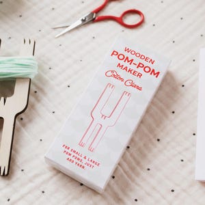 May include: A white box with red text and illustrations for a wooden pom-pom maker. The box reads "WOODEN POM-POM MAKER Cotton Clara" and "FOR SMALL & LARGE POM POMS, JUST ADD YARN." Red scissors and a wooden pom-pom maker are also visible.