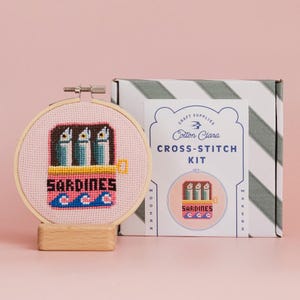 May include: A cross-stitch kit with a design of sardines in a can. The embroidery hoop is displayed on a wooden stand. The kit includes a box with the text "Cotton Clara Cross-Stitch Kit". The design uses pink, blue, and yellow colours.