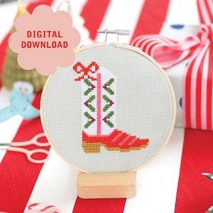 May include: A cross-stitch embroidery hoop featuring a red, white, and green cowboy boot design. The boot is decorated with a bow and festive accents. The text "DIGITAL DOWNLOAD" is in a pink cloud-shaped speech bubble.