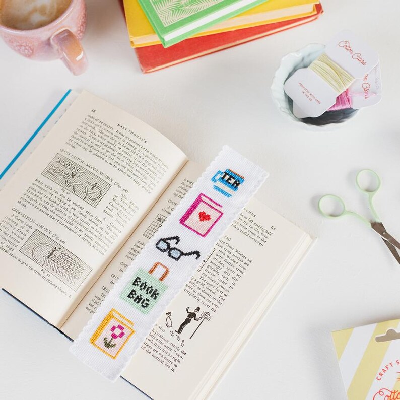 May include: White bookmark with colorful cross-stitch embroidery of a book bag, coffee cup, glasses, flower, and heart. The bookmark is laying on top of an open book.