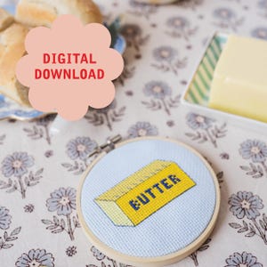 Pode incluir: Um bastidor de bordado em ponto cruz com um desenho de manteiga amarela e a palavra "BUTTER" em azul. O bastidor está sobre uma toalha de mesa floral. Um gráfico de nuvem rosa com "DIGITAL DOWNLOAD" está no canto superior esquerdo.