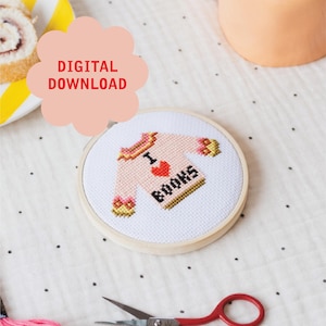 May include: A cross-stitch embroidery hoop featuring a pink sweater design with the words "I BOOKS" and a red heart. The words "DIGITAL DOWNLOAD" are in a pink cloud-shaped graphic. A pair of red-handled scissors is in the foreground.
