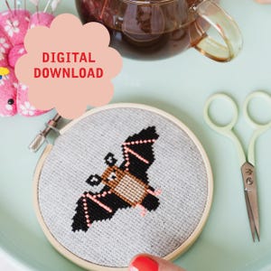Può includere: Un telaio da ricamo a punto croce con un disegno di pipistrello nero e marrone con accenti rosa. Il testo "DIGITAL DOWNLOAD" è in una grafica a forma di nuvola rosa. Una tazza di tè e delle forbici sono anche nell'immagine.
