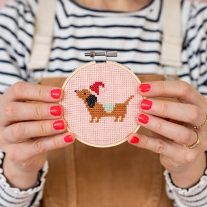 May include: A pink and brown cross-stitch embroidery hoop with a dachshund wearing a Santa hat. The hoop is held by a person's hands.