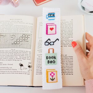 May include: White bookmark with colorful cross-stitch embroidery of a teacup, book, glasses, shopping bag, and flower. The bookmark is on a book with text about cross-stitch.