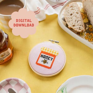 May include: A pink and white cross-stitch embroidery hoop with a honey jar design. The jar is orange with a white and yellow checkered lid and the word "honey" in black letters. A small bee is below the word "honey".