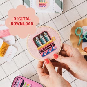 May include: A cross-stitch embroidery hoop featuring a sardine can design with the word "SARDINES" and blue waves. The text "DIGITAL DOWNLOAD" is in a pink cloud shape. The hoop is held by a person with red nail polish.