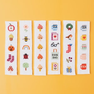 May include: A collection of six white cross-stitch bookmarks with various designs. The designs include an apple, a pumpkin, a bee, a rainbow, mushrooms, a book, a bird, a wreath, and the word "TEA".
