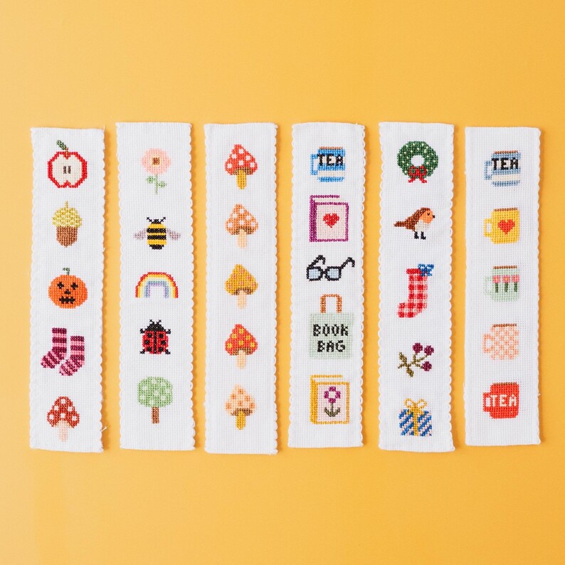 May include: A collection of six white cross-stitch bookmarks with various designs. The designs include an apple, a pumpkin, a bee, a rainbow, mushrooms, a book, a bird, a wreath, and the word "TEA".