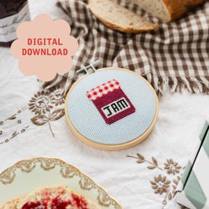 Puede incluir: Un aro de bordado de punto de cruz con fondo blanco y un tarro de mermelada a cuadros rojos y blancos con la palabra "JAM" en letras negras.