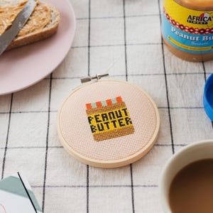 May include: A close-up of a small, round, wooden embroidery hoop with a pink fabric background. The fabric has a cross-stitch design of a peanut butter jar with the words "Peanut Butter" in black letters.