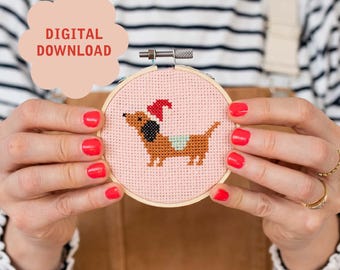 May include: A pink embroidery hoop with a cross-stitch design of a dachshund wearing a Santa hat. The dachshund is brown with a light blue and white saddle.