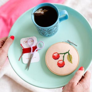 May include: A teal tray with a blue mug filled with black tea, a small spool of red embroidery floss, a pair of purple scissors, and a small embroidery hoop with a cross-stitch design of two red cherries on a green stem.