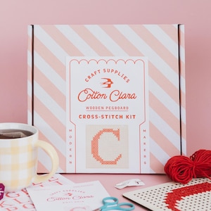 Può includere: Una scatola a righe rosa e bianche con il testo "CRAFT SUPPLIES Cotton Clara WOODEN PEGBOARD CROSS-STITCH KIT" sul davanti. La scatola contiene un kit per ricamo a punto croce con una C rossa su una tela bianca, filo rosso, un telaio in legno e forbici.