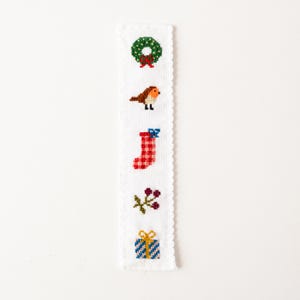 May include: A white, rectangular bookmark with a scalloped edge, featuring cross-stitch designs of a green wreath, a bird, a red stocking, berries, and a wrapped gift. The designs are in various colors, including red, green, blue, and yellow.