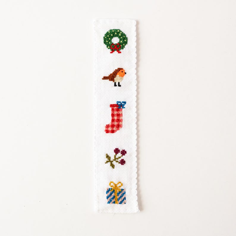 May include: A white, rectangular bookmark with a scalloped edge, featuring cross-stitch designs of a green wreath, a bird, a red stocking, berries, and a wrapped gift. The designs are in various colors, including red, green, blue, and yellow.