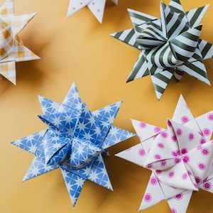 May include: A collection of paper star decorations in various patterns. The stars display designs such as blue and white geometric shapes, green and white stripes, and pink polka dots. The background is a plain yellow.