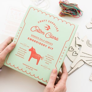 Può includere: Una scatola verde con il testo "Craft Supplies Cotton Clara Wooden Dala Horses Embroidery Kit" e il testo "Everything you need to make this banner is included in this kit. One stitch at a time." La scatola è tenuta da due mani. La scatola è su una superficie bianca con un ritaglio di cavallo di legno.