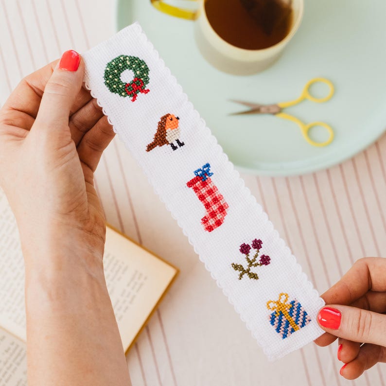 May include: White cross-stitch bookmark with a scalloped edge, featuring Christmas-themed designs. The designs include a wreath, bird, stocking, berries, and a wrapped gift. The bookmark is held by a person.