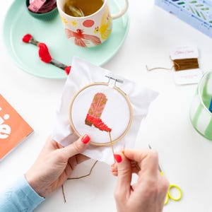 May include: A close-up of a person cross-stitching a red and pink cowboy boot on white Aida cloth in a wooden embroidery hoop. The hoop is on a white surface with a spool of brown embroidery floss and a pair of scissors.