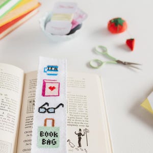 May include: A white bookmark with a cross-stitch design featuring a cup of tea, a book with a heart, a pair of glasses, and a green book bag with the text "Book Bag".