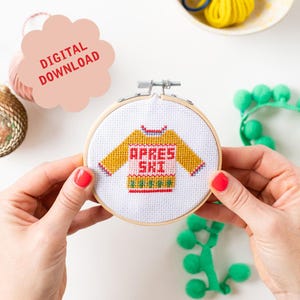 May include: A white embroidery hoop with a cross-stitch design of a yellow sweater with the text "Apres Ski" in red and white. The sweater has a green and white snowflake pattern on the bottom.