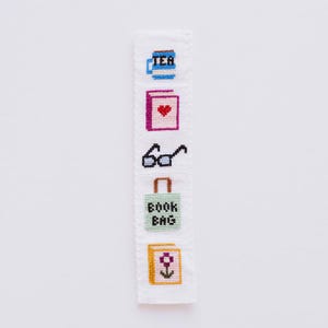 May include: A white bookmark with a stitched design featuring a cup of tea, a book with a heart, glasses, a book bag, and a book with a flower. The bookmark has a scalloped edge.