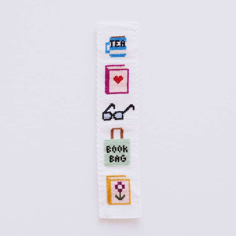 May include: A white bookmark with a stitched design featuring a cup of tea, a book with a heart, glasses, a book bag, and a book with a flower. The bookmark has a scalloped edge.