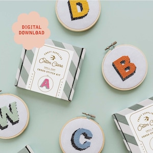 May include: A set of five cross-stitch kits featuring the letters A, B, C, D, and W. Each kit includes a small wooden hoop, embroidery floss, and a pattern for stitching the letter. The kits are packaged in a white box with a green and white striped design. The box has the text "Cotton Clara" and "Small Hoop Cross-Stitch Kit" printed on it.