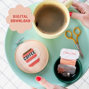 Peut inclure: Un gros plan d'un cerceau de broderie en point de croix avec un motif de tasse de café rouge. Le cerceau est sur un plateau turquoise avec une tasse de café, des ciseaux et une bobine de fil à broder rose.