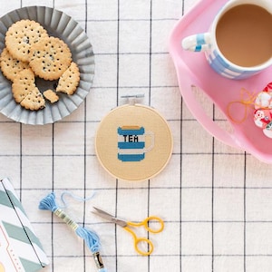 Peut inclure: Un gros plan d'un cerceau de broderie en point de croix avec un motif de tasse à thé rayée bleu et blanc. La tasse à thé porte l'inscription "TEA". Le cerceau est sur une surface à carreaux blancs et roses.