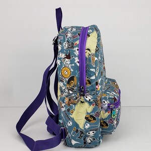 May include: A blue and green backpack with a purple zipper and straps. The backpack is covered in a pattern of characters from the Nightmare Before Christmas.