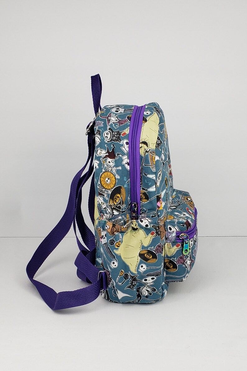 May include: A blue and green backpack with a purple zipper and straps. The backpack is covered in a pattern of characters from the Nightmare Before Christmas.
